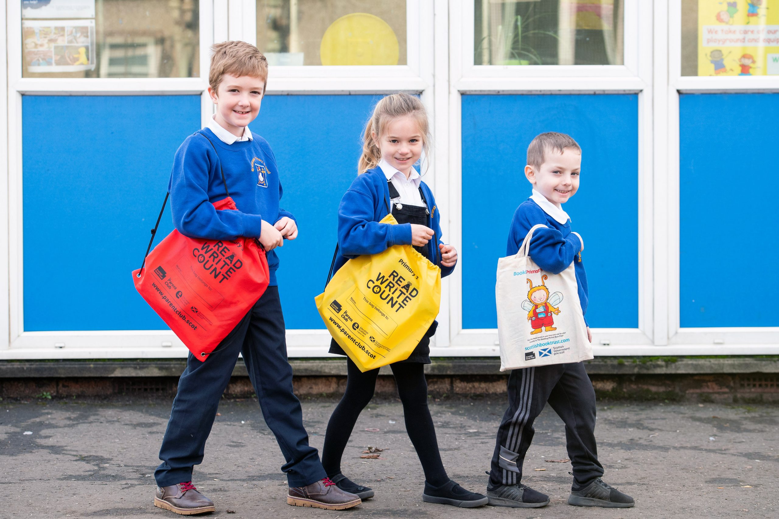 Three primary school children carrying book bags stand outside their school