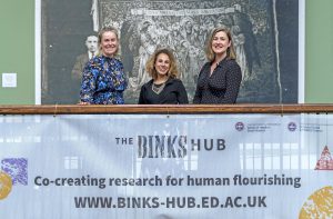 Our three co-directors stand on a balcony above a sign reading 'The Binks Hub'