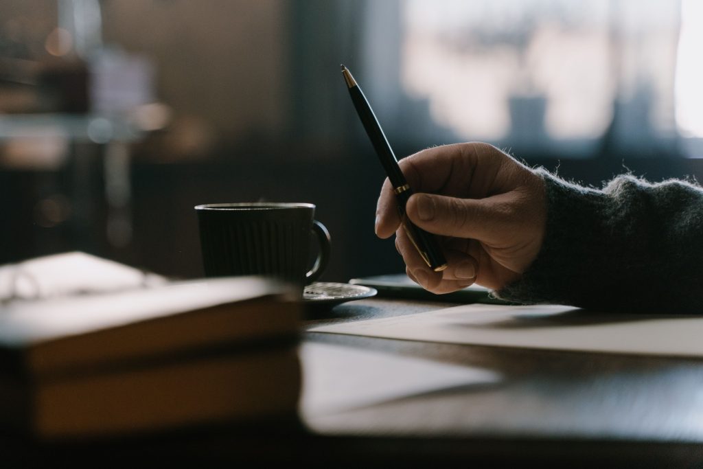 Decorative image: close-up of a hand holding a pen over paper, book in the foreground.